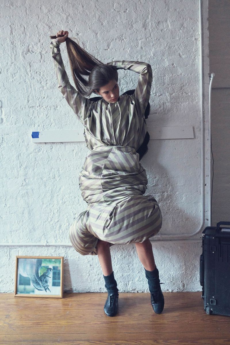 Woman in a sculptural striped dress stands in a room, holding her hair up with both hands, with a picture frame and luggage nearby.