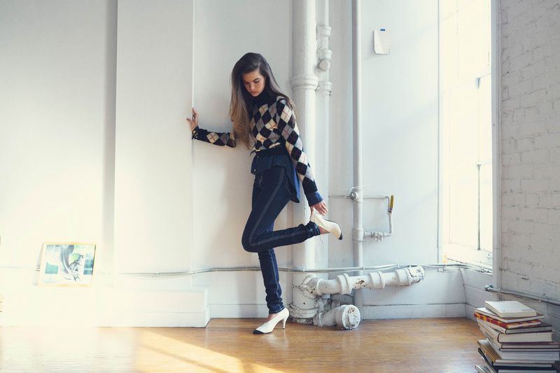 Model in argyle sweater and dark jeans leaning against a white pillar in a bright room with books stacked on the floor