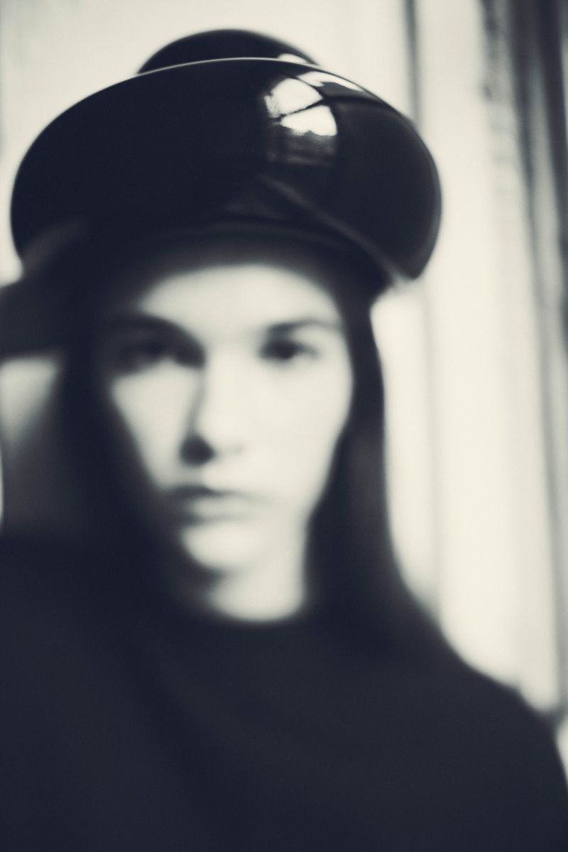 Portrait of a blurred woman with long hair wearing a glossy black hat and top, set against a softly lit white background.