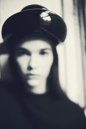 Portrait of a blurred woman with long hair wearing a glossy black hat and top, set against a softly lit white background.