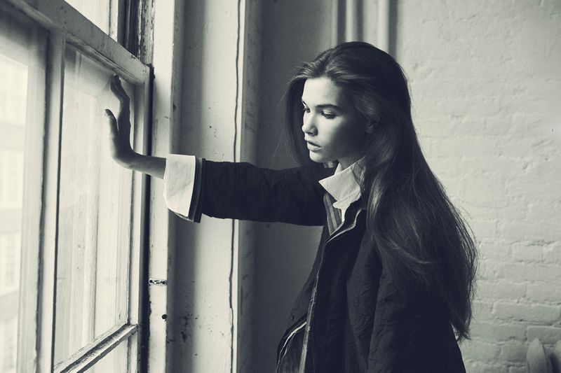 Monochrome portrait of a young woman in a black jacket standing by a window, with long dark hair and a pensive expression.