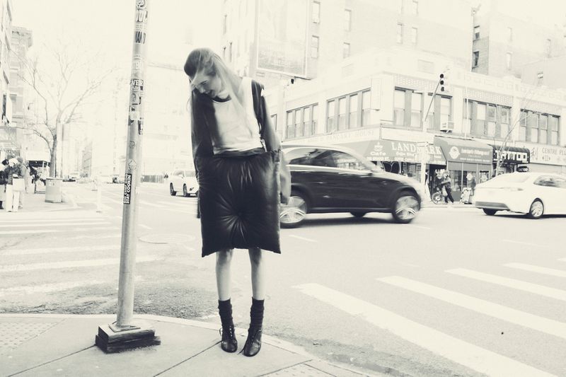 Monochrome full shot of a woman in avant-garde fashion posing on a city street corner with blurred traffic in the background.