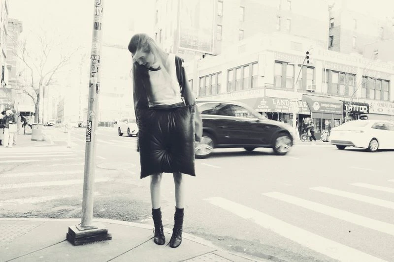 Monochrome full shot of a woman in avant-garde fashion posing on a city street corner with blurred traffic in the background.
