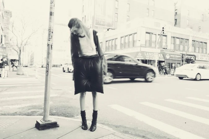 Monochrome full shot of a woman in avant-garde fashion posing on a city street corner with blurred traffic in the background.