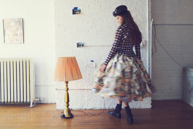 Fashionable woman in ornate dress poses indoors by a vintage floor lamp, showcasing unique style and artistic design in a bright space.