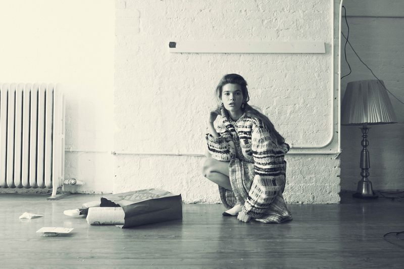 A woman in a patterned sweater dress crouching indoors near a radiator and discarded paper, captured in a black and white aesthetic.