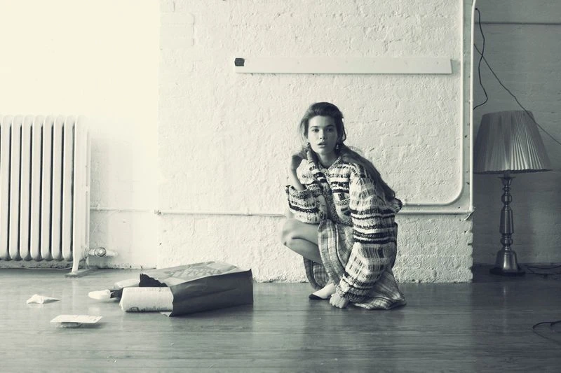 A woman in a patterned sweater dress crouching indoors near a radiator and discarded paper, captured in a black and white aesthetic.