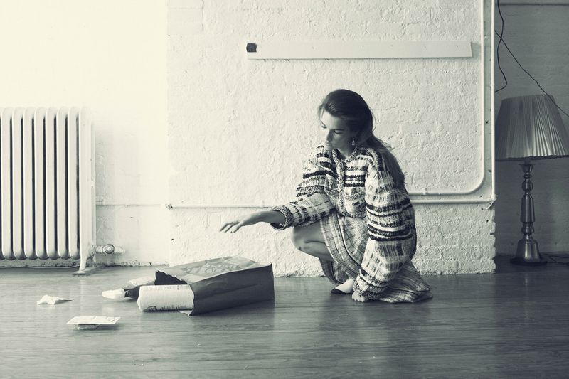 Woman in patterned dress crouches with hand extended toward papers spilling from a brown paper bag on a wood floor in a room.