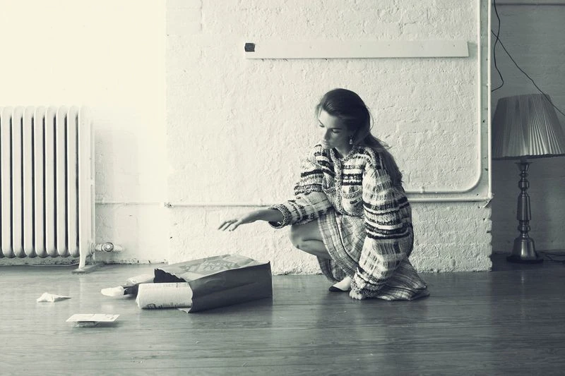 Woman in patterned dress crouches with hand extended toward papers spilling from a brown paper bag on a wood floor in a room.
