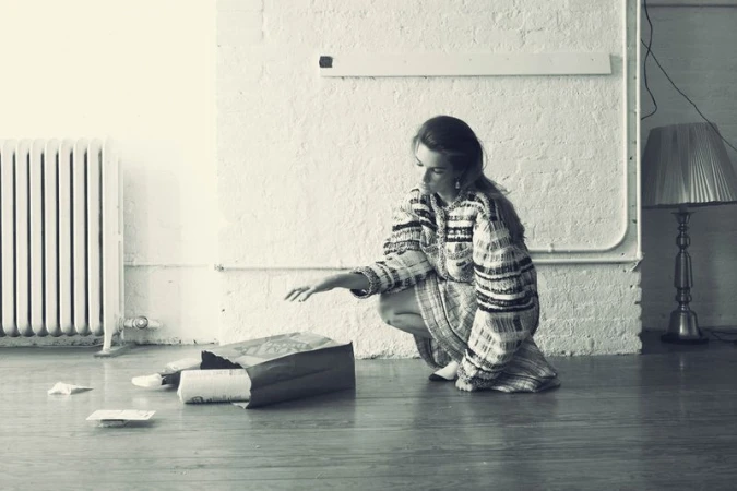 Woman in patterned dress crouches with hand extended toward papers spilling from a brown paper bag on a wood floor in a room.