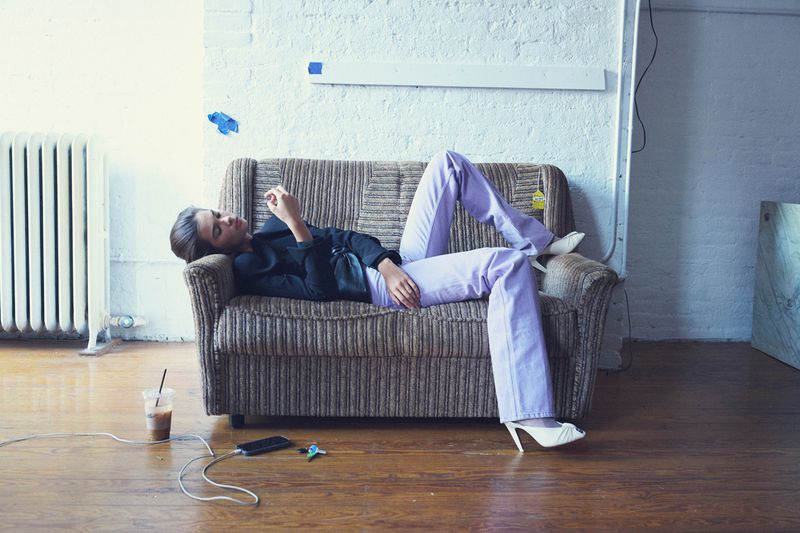 Woman lounges on a couch with lavender pants and white heels, engrossed in her phone, with a drink and charging cable nearby.