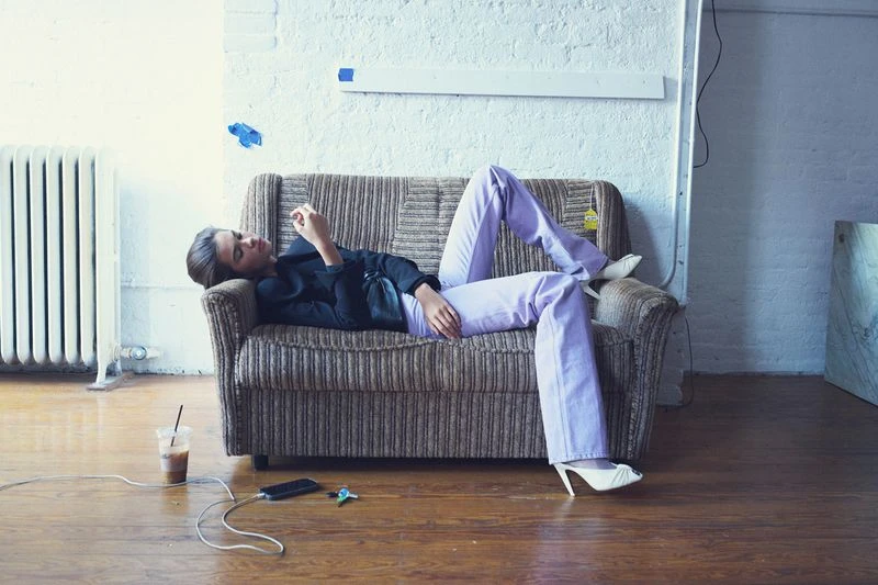 Woman lounges on a couch with lavender pants and white heels, engrossed in her phone, with a drink and charging cable nearby.