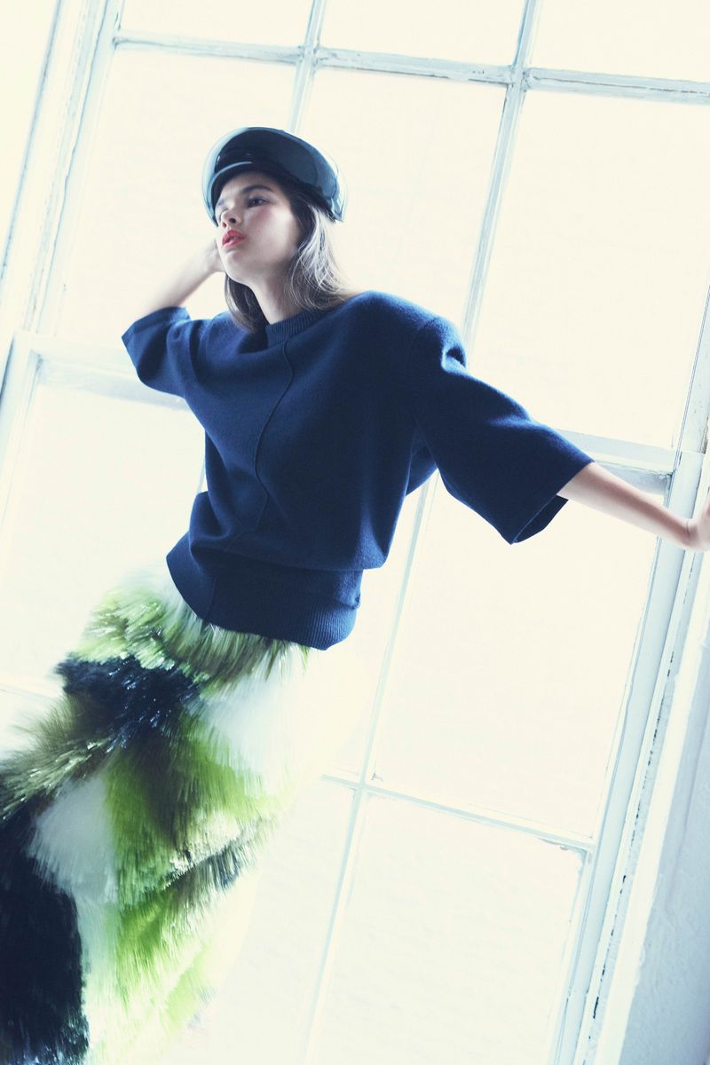 A young woman in a navy sweater and fuzzy green and white skirt poses next to a window with bright, diffused light. She wears a navy cap.