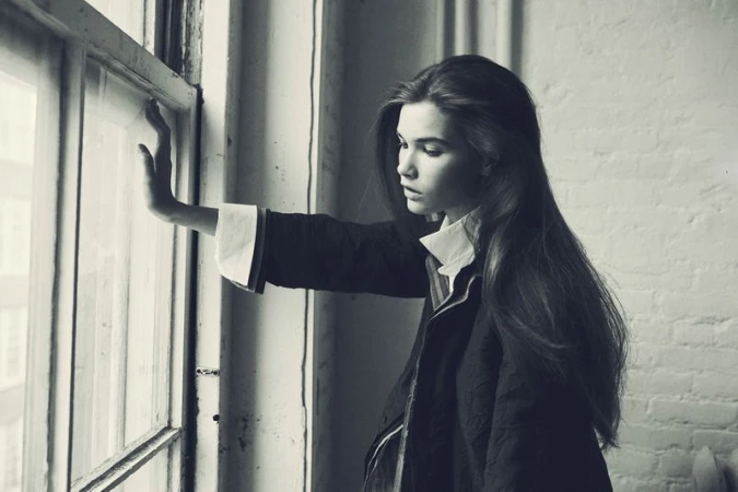 A young female model with long dark hair leaning against a window frame in a monochromatic editorial portrait.