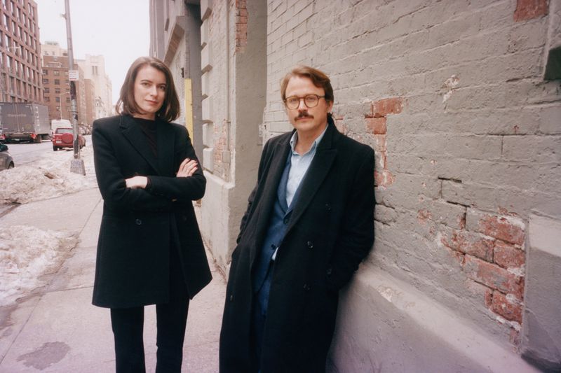 A fashionable couple poses against a brick wall on a city sidewalk, captured in natural light, evoking a candid, urban editorial style.