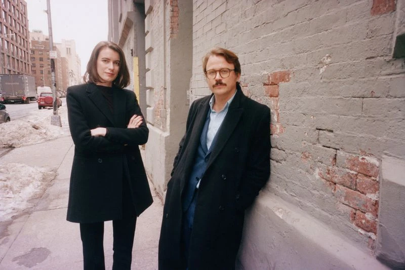 A fashionable couple poses against a brick wall on a city sidewalk, captured in natural light, evoking a candid, urban editorial style.