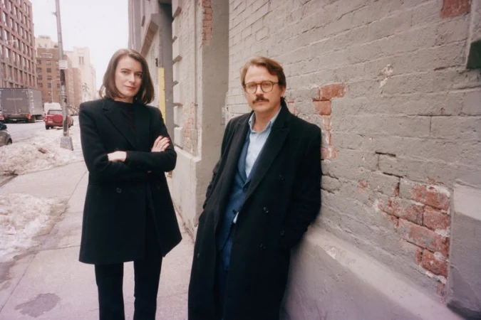 A fashionable couple poses against a brick wall on a city sidewalk, captured in natural light, evoking a candid, urban editorial style.