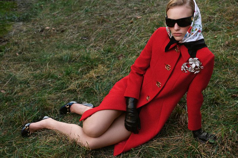 Model wearing a bright red coat, headscarf, and sunglasses reclines on a grass-covered outdoor terrain.