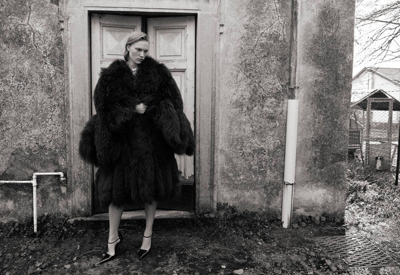 Woman in oversized fur coat and heels stands in front of a weathered building. Black and white fashion shot with an aged appearance.