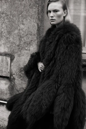 Monochrome portrait of a blond adult female fashion model wearing a large dark furry coat, with a stone wall background.