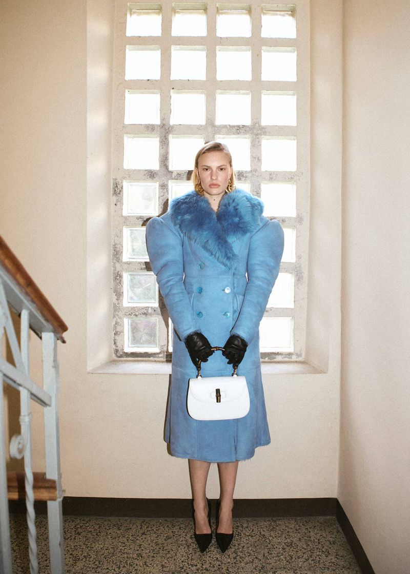 A woman in a blue coat poses with a white purse inside a building with a gridded window, exuding a stylish and fashionable look.