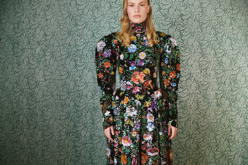 A blonde female fashion model wears a long floral dress in a studio with a patterned backdrop for a professional fashion editorial.