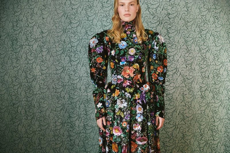 A blonde female fashion model wears a long floral dress in a studio with a patterned backdrop for a professional fashion editorial.