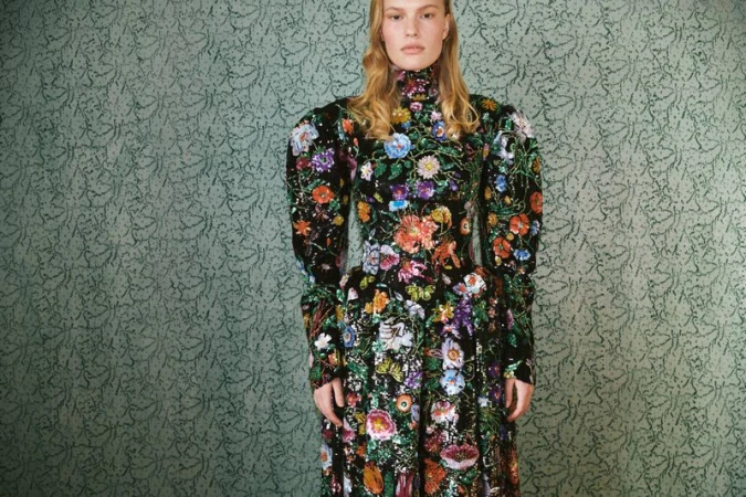 A blonde female fashion model wears a long floral dress in a studio with a patterned backdrop for a professional fashion editorial.