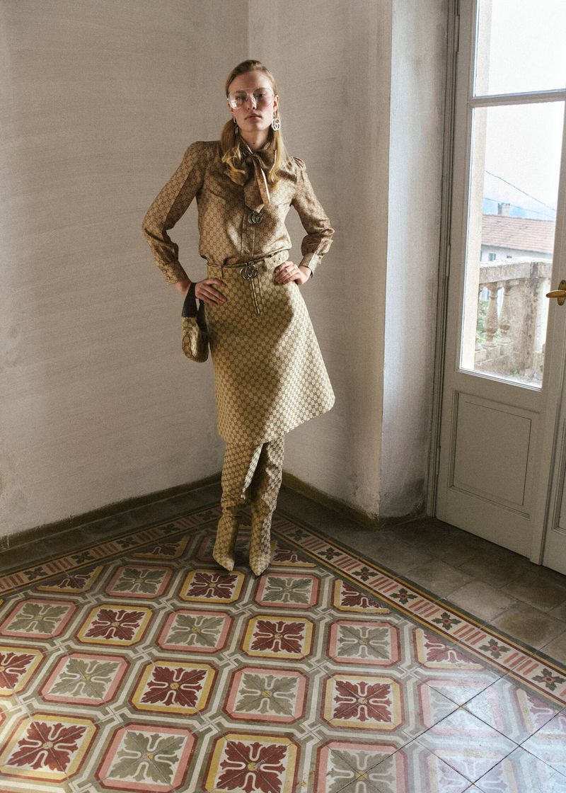 Model wearing patterned beige blouse and skirt standing on tiled floor near door for Muse Magazine editorial.