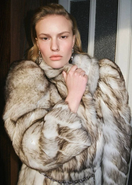 Elegant adult woman in a luxurious fur coat poses indoors, showcasing fashion style with door background and soft lighting.