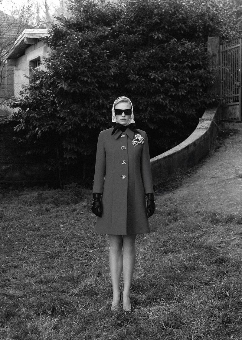 Monochrome fashion shot of a woman in a red coat, headscarf, and sunglasses standing in a grassy outdoor setting with a house in the background.