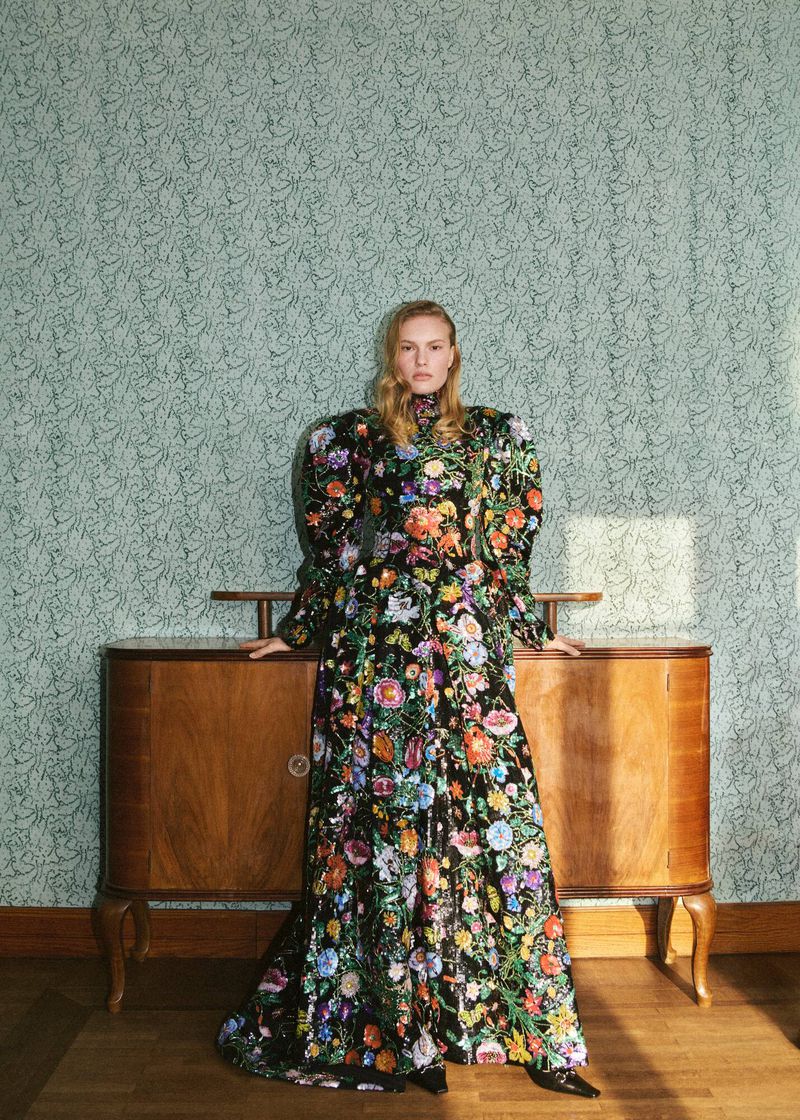 A full shot of a blond adult woman posing in a floral black dress in front of a wood credenza against a floral wallpaper background.