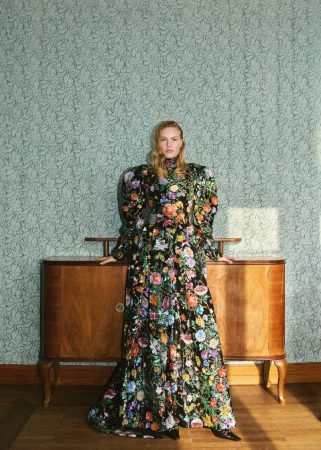 A full shot of a blond adult woman posing in a floral black dress in front of a wood credenza against a floral wallpaper background.