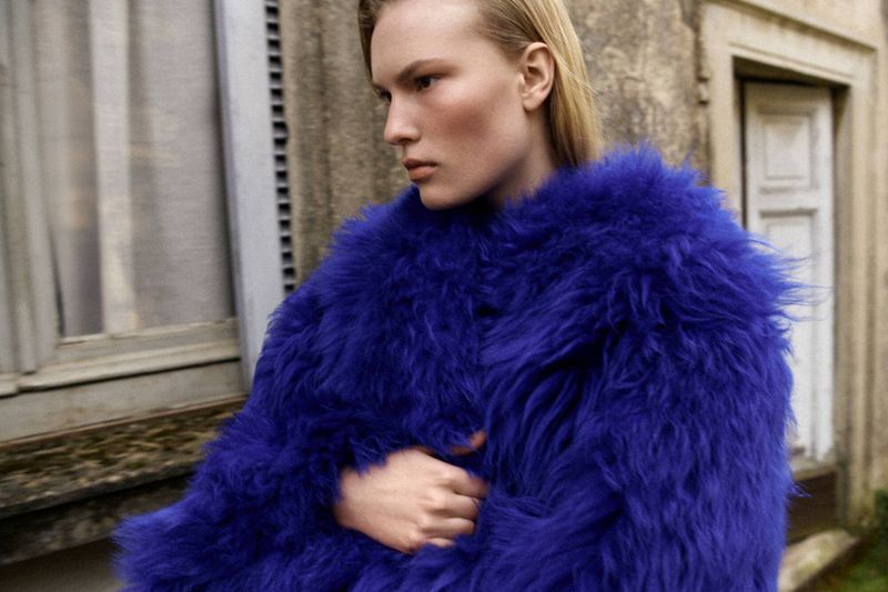 Close-up shot of a blonde-haired woman wearing a blue fur coat, posing in front of a weathered stone building with windows and a door.
