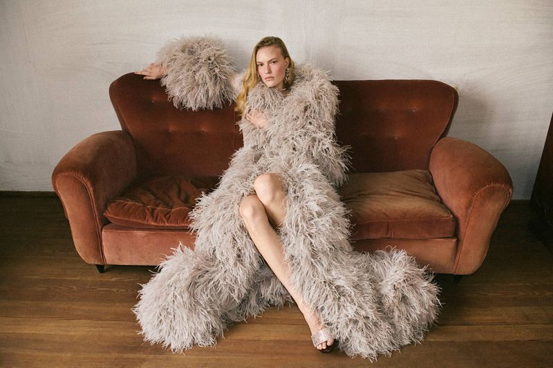 Adult woman sits on a vintage couch wearing a long feathered robe and heels in a fashion editorial shot, indoors with soft lighting.