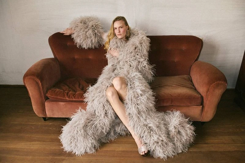 Adult woman sits on a vintage couch wearing a long feathered robe and heels in a fashion editorial shot, indoors with soft lighting.
