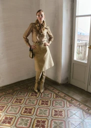 Model wearing patterned beige blouse and skirt standing on tiled floor near door for Muse Magazine editorial.