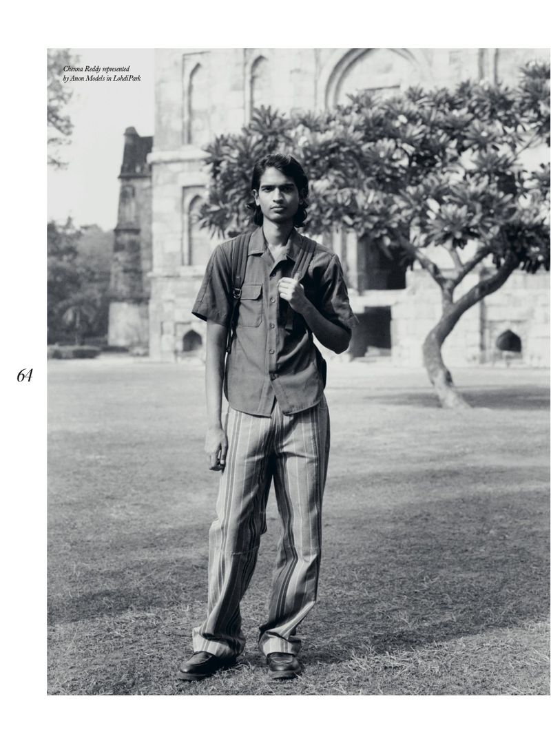 Portrait of a young adult male wearing a button-down shirt and striped trousers standing in a park with an old building behind him.