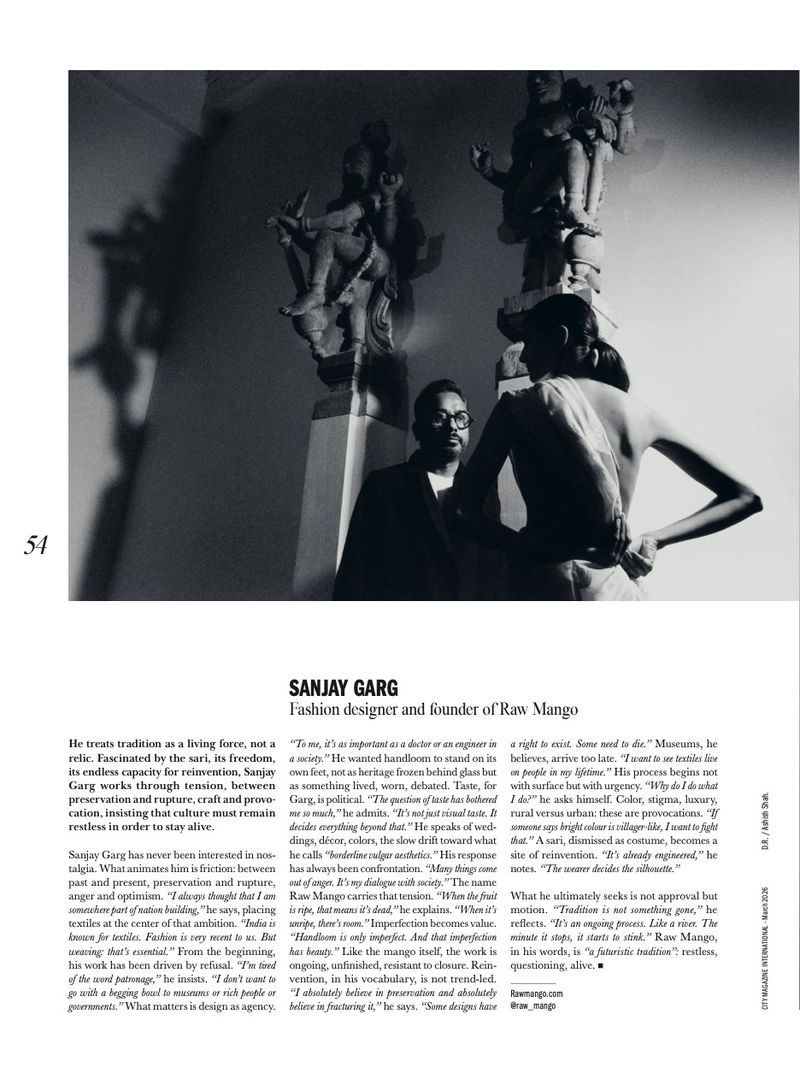 Monochrome portrait of Sanjay Garg, fashion designer, standing in front of ornate sculptures, wearing glasses and a dark suit, editorial style.
