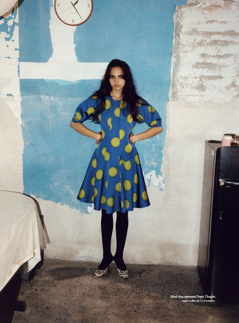 A full shot of a young woman in a blue polka-dot dress with dark tights and zebra-print shoes, standing in a room with a distressed wall.