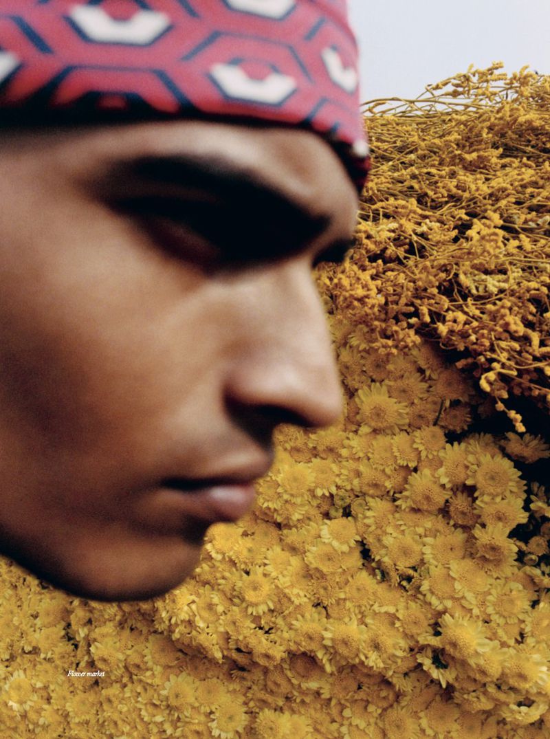 Close-up of a person with yellow flower field in the background. The model wears a red and white geometric patterned head wrap.