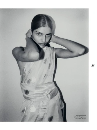 Monochrome portrait of a South Asian woman in a sari, hands behind head, looking at the camera in a simple indoor setting.