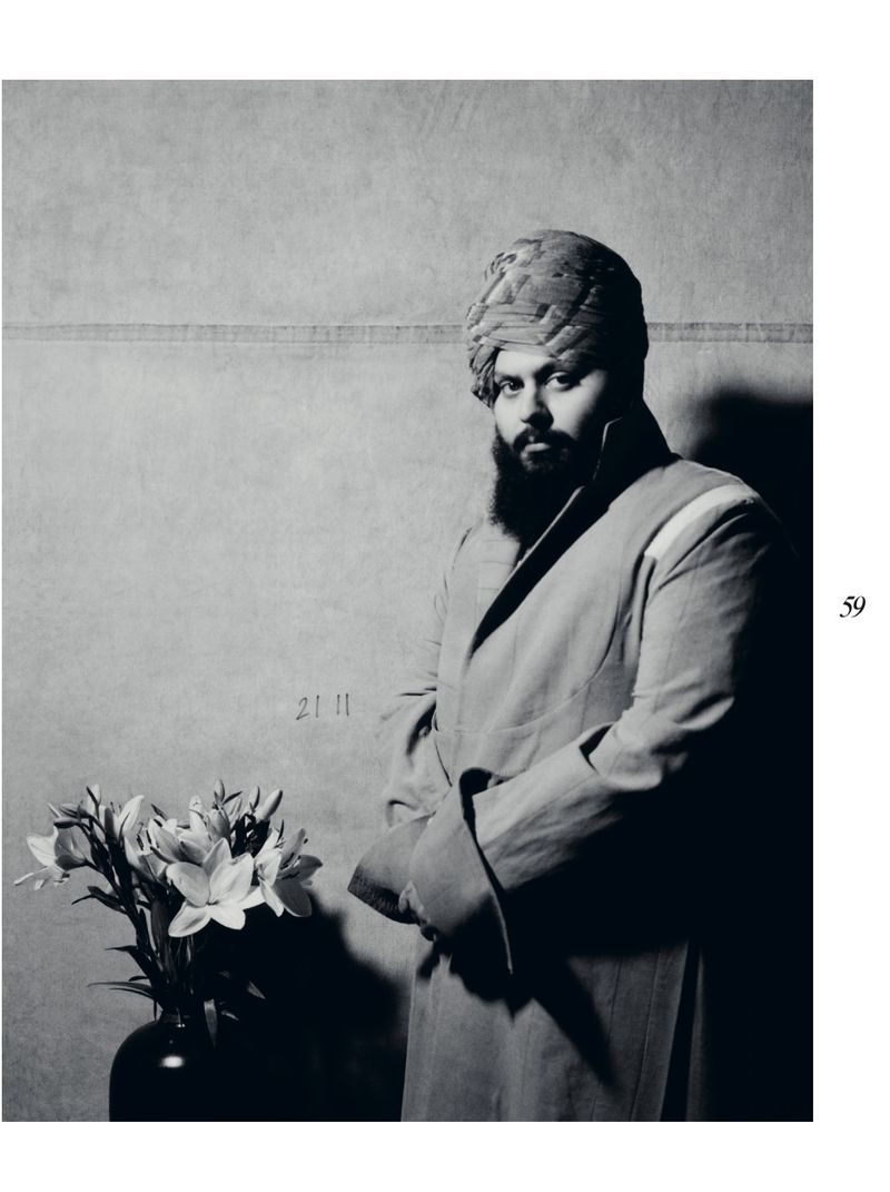 Monochrome portrait of a turbaned adult male in a long coat indoors with lilies, in front of a neutral-toned backdrop and soft lighting.