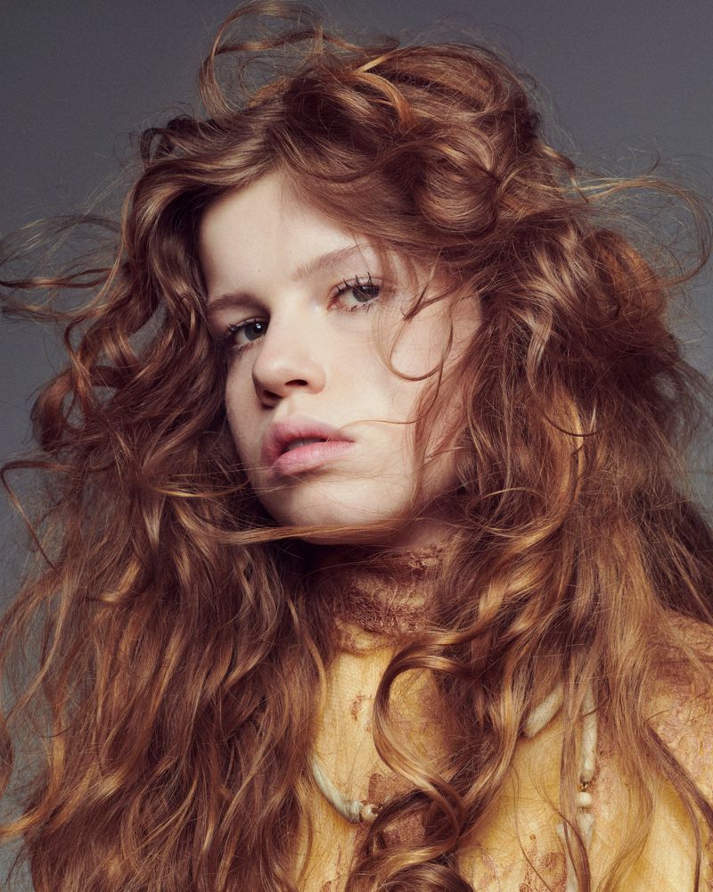Close-up portrait of a young woman with long, curly, reddish-brown hair, fair skin, and a neutral facial expression indoors against a grey backdrop.