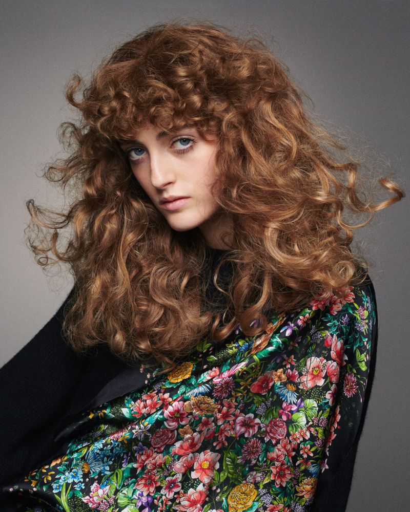 Portrait of a Caucasian woman with curly auburn hair wearing a floral-print scarf and black top, posing in a studio setting.
