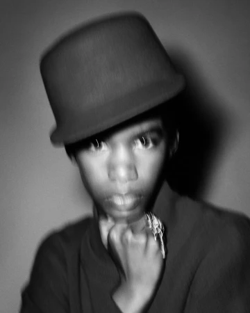 Portrait of a person wearing a dark bowler hat, hands clasped near chin, captured with a soft motion blur effect in black and white.