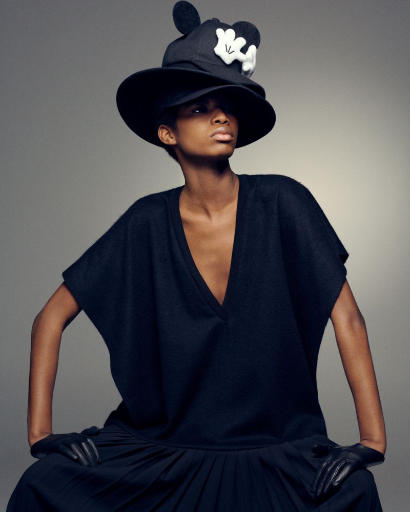 A fashion model wearing a black hat with white glove detail and a black v-neck top with pleated skirt against a grey background.