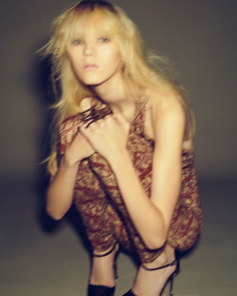 Young woman with long blonde hair, wearing a patterned jumpsuit and strappy heels, crouching indoors with soft studio lighting.