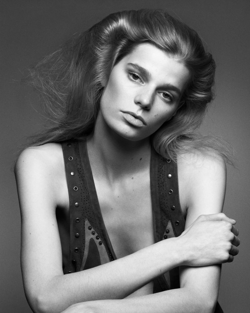Black and white portrait of a young woman with voluminous blonde hair, wearing a studded vest, in a studio setting.
