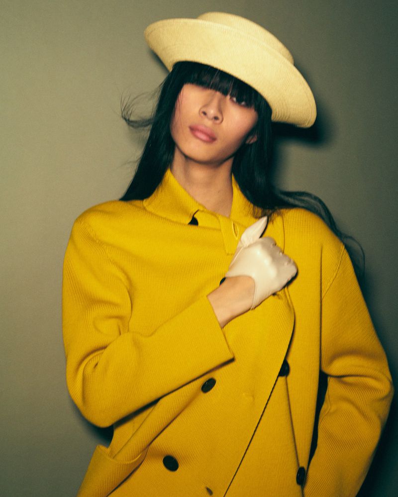 A fashionable young Asian woman with a modern hairstyle is wearing a yellow jacket, white gloves and hat while posing in an indoor setting.